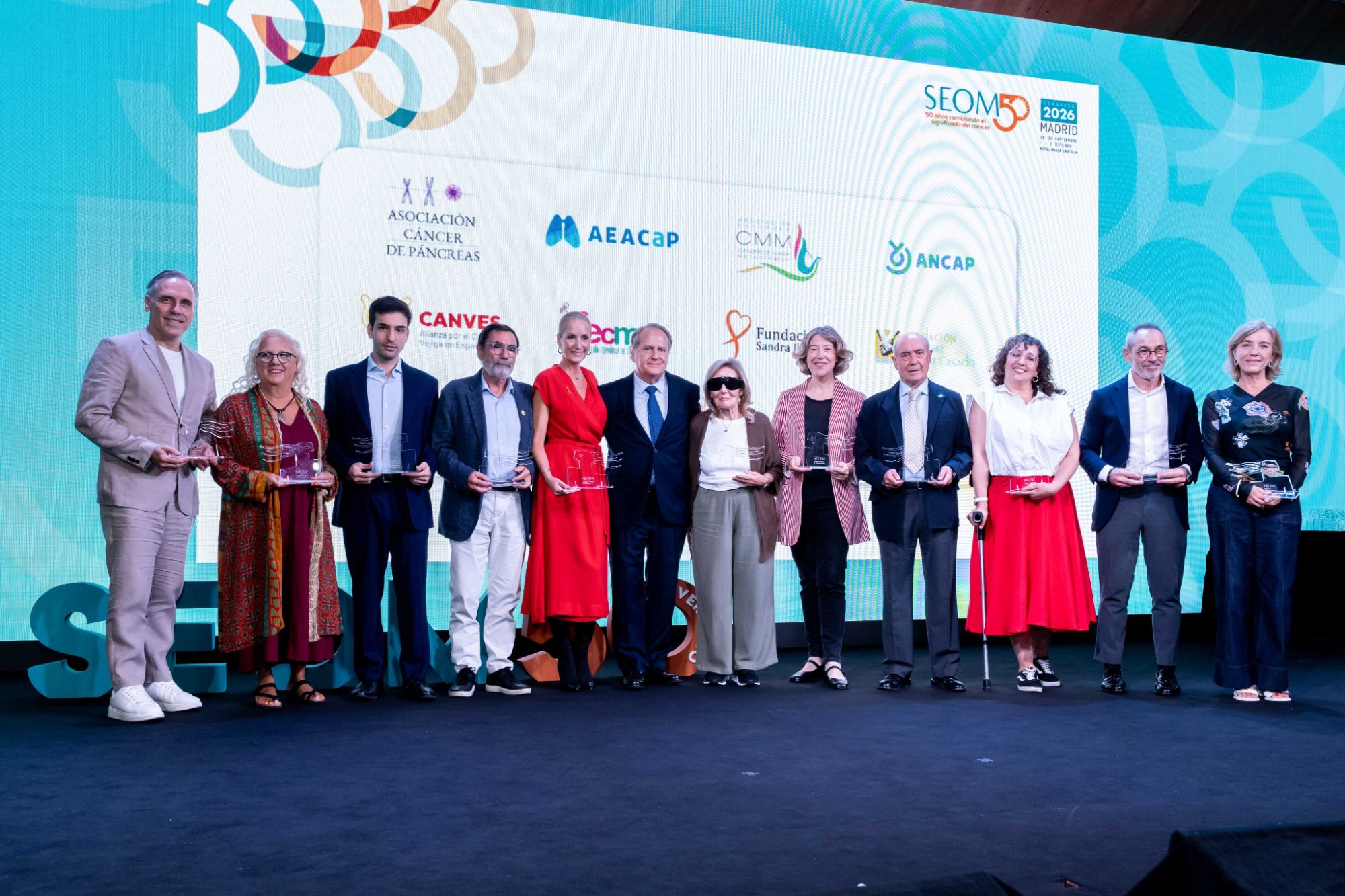 Entidades y personas reconocidas en la Gala de Homenaje a Pacientes de Cáncer y Cuidadores, con sus galardones, ante el cartel del 50 aniversario de SEOM